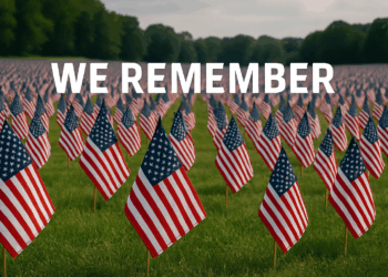 A grassy field with small American flags arranged in rows the whole length of the field. Over the flags, the text We Remember is visible.